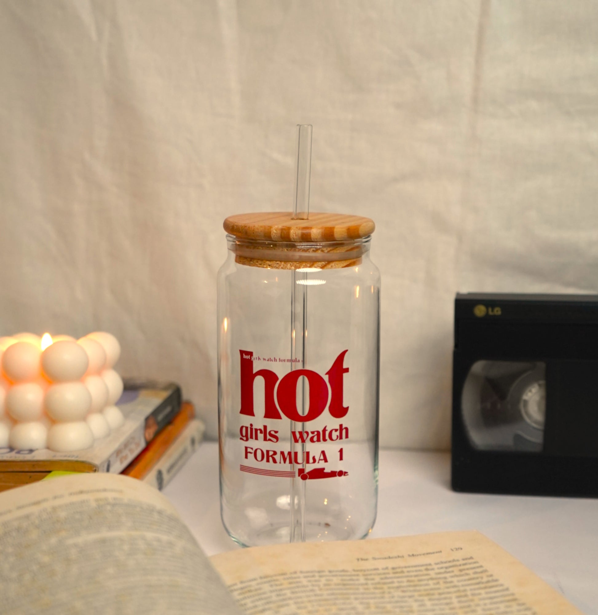 Clear glass tumbler with wooden lid and red text on a surface with books and a candle.