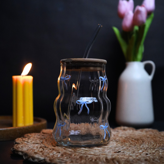 Glass wavy tumbler with floral design on a woven mat, candle, and vase with flowers in the background.